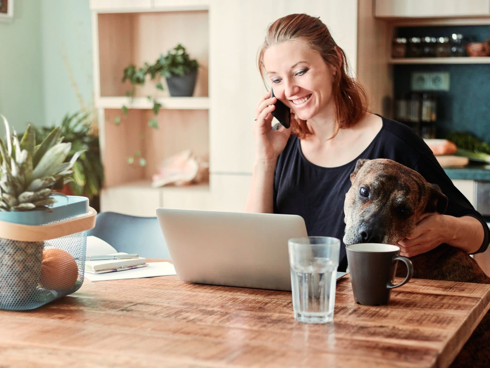 A woman smiling on the phone at her kitchen table, her dog beside her.