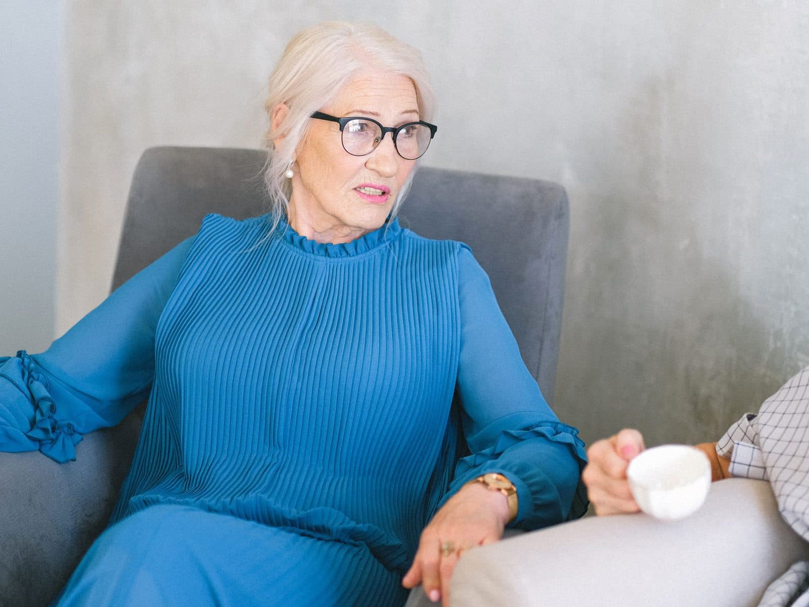 An older woman in a blue dress chatting warmly from her armchair at home.