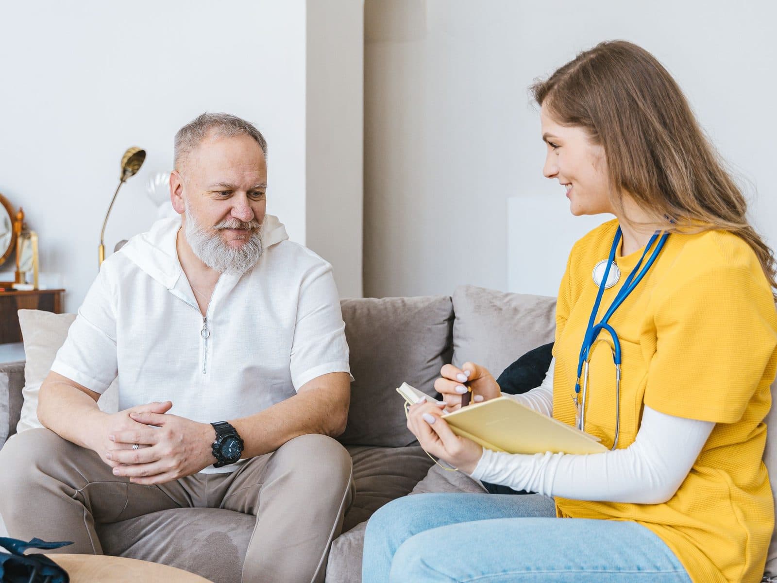 A nurse with a notepad listening to an older man on his sofa at home.