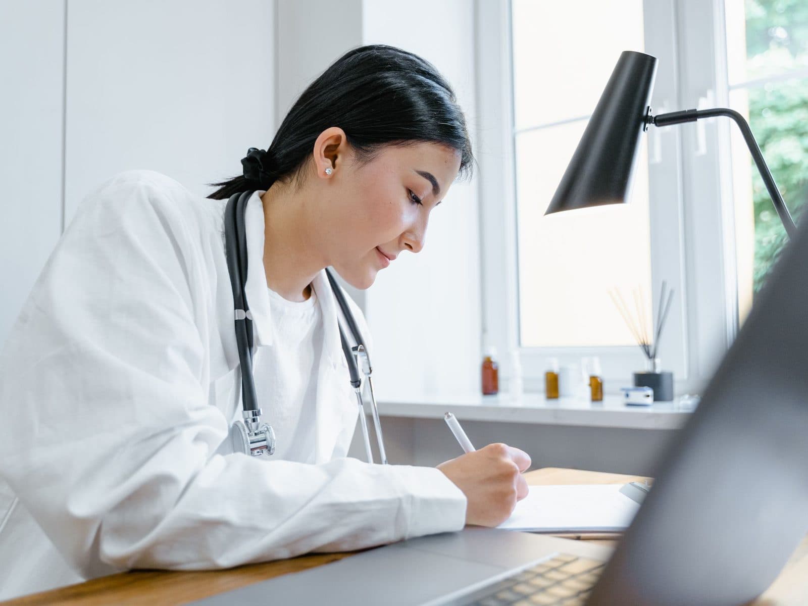 A doctor writing a referral at her desk by a bright window.